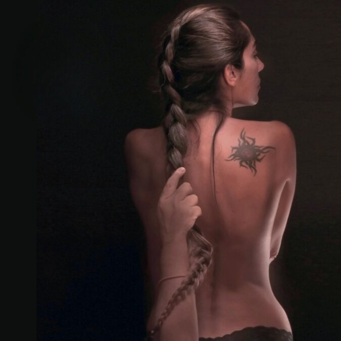 Una mujer de espaldas, desnuda de cintura para arriba, con una trenza larga sostenida por una mano. En su hombro derecho se observa un tatuaje negro con forma de sol. La imagen transmite una atmósfera de sensualidad y misterio.