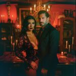 Una pareja elegante, el hombre en traje negro y la mujer en un vestido negro escotado, posando en un ambiente decorado con velas y un candelabro