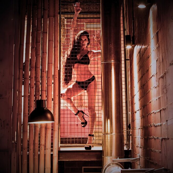 Mujer en lencería negra trepando dentro de una celda iluminada en un espacio industrial. Promoción de la Fiesta de la Liberación en Open-Minded Valencia.