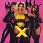 Grupo de mujeres con trajes de cuero y látex, posando con confianza frente a un fondo rosa con una gran “X” amarilla