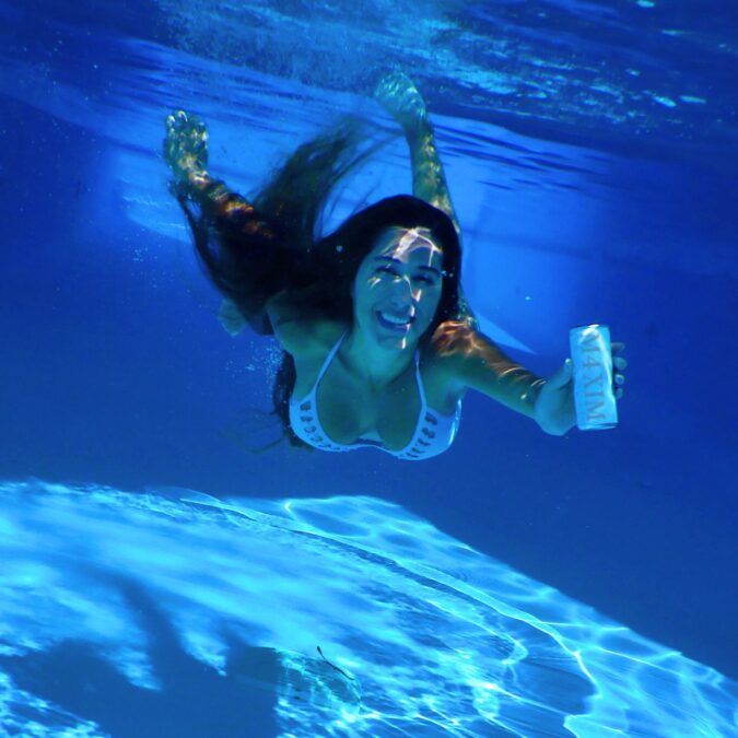 Mujer nadando bajo el agua con una sonrisa, sosteniendo una bebida en la mano, en un ambiente de luz azul.