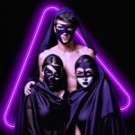 Hombre y dos mujeres con máscaras y capas negras, iluminados por un triángulo de neón morado en el fondo