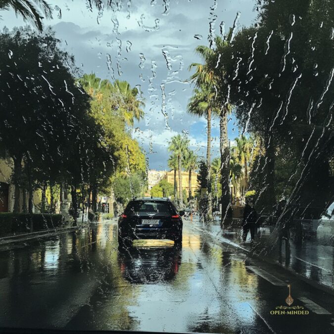Vista de una calle en Valencia después de la lluvia, con gotas en el parabrisas y palmeras a los lados de la carretera. Logo de OPEN-MINDED en la esquina inferior derecha