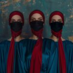 Tres mujeres con turbantes rojos y máscaras negras, vestidas con túnicas azules, frente a un fondo texturizado en tonos dorados y azules.